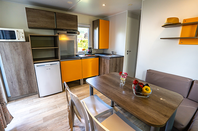 The kitchen and living room area with a table, chairs and sofa bed for 2 people of the Brand mobile home for 6 people, for rent at the Médiéval campsite near Colmar